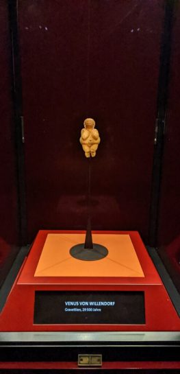 The Venus of Willendorf sitting in her glass case on a black background. The museum label reads "Venus von Willendorf."